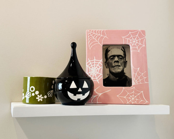 Halloween-themed decor items including a black vase with a pumpkin face, a green mug with white floral design, and a pink frame with a spider web pattern on a shelf.