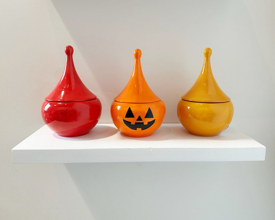 Three ceramic jars in red, orange, and yellow on a white shelf against a light gray background.