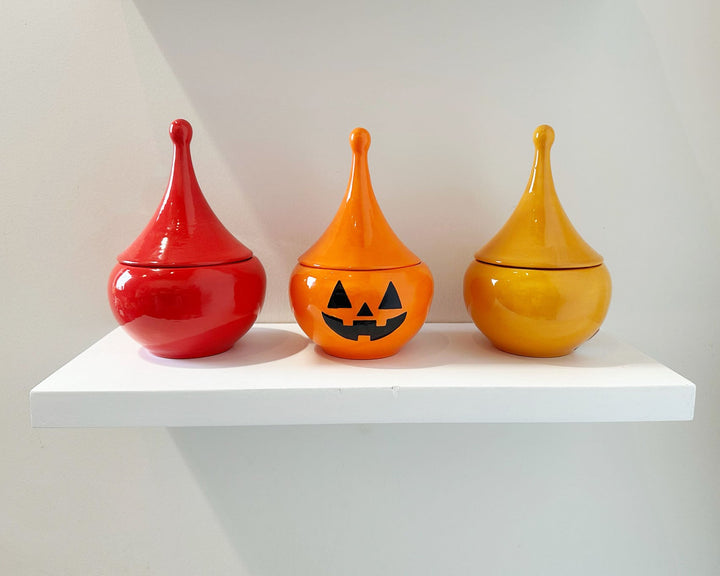 Three ceramic jars in red, orange, and yellow on a white shelf against a light gray background.