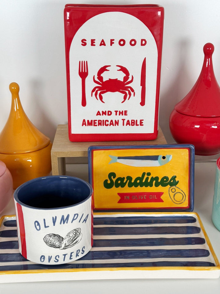 Decorative items including a sign with a crab and text, a cup with 'Olympia Oysters' text, and a small box with 'Sardines' text on a striped surface.