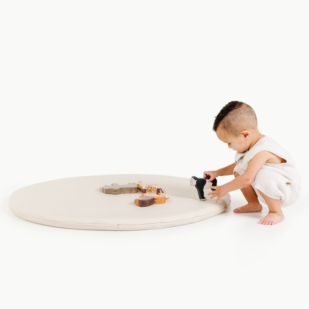 Cream round padded play mat with a toddler kneeling to play with wooden toys.