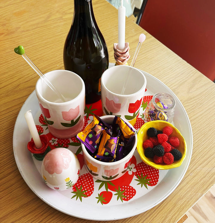 Pink tulip cups with stir sticks and one filled with candy on a strawberry platter beside berries and a bottle