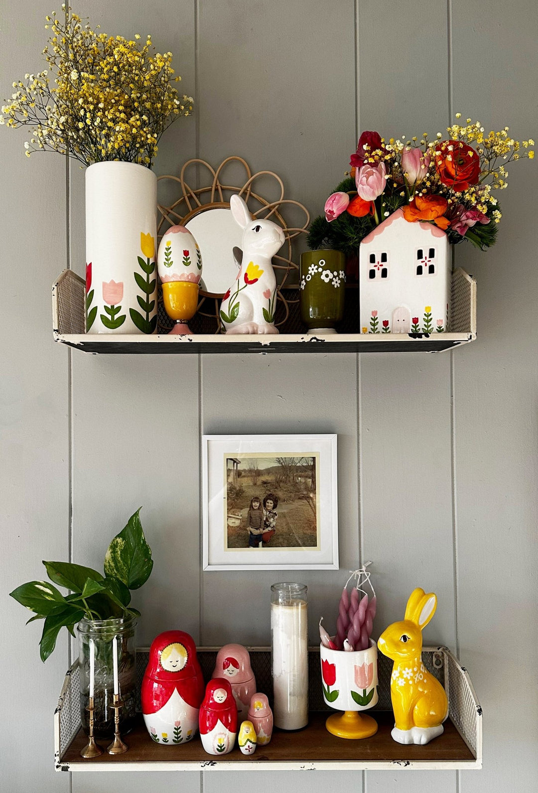 Two wall shelves styled with a tulip cup, a tulip house vase of flowers, nesting dolls, and a yellow bunny