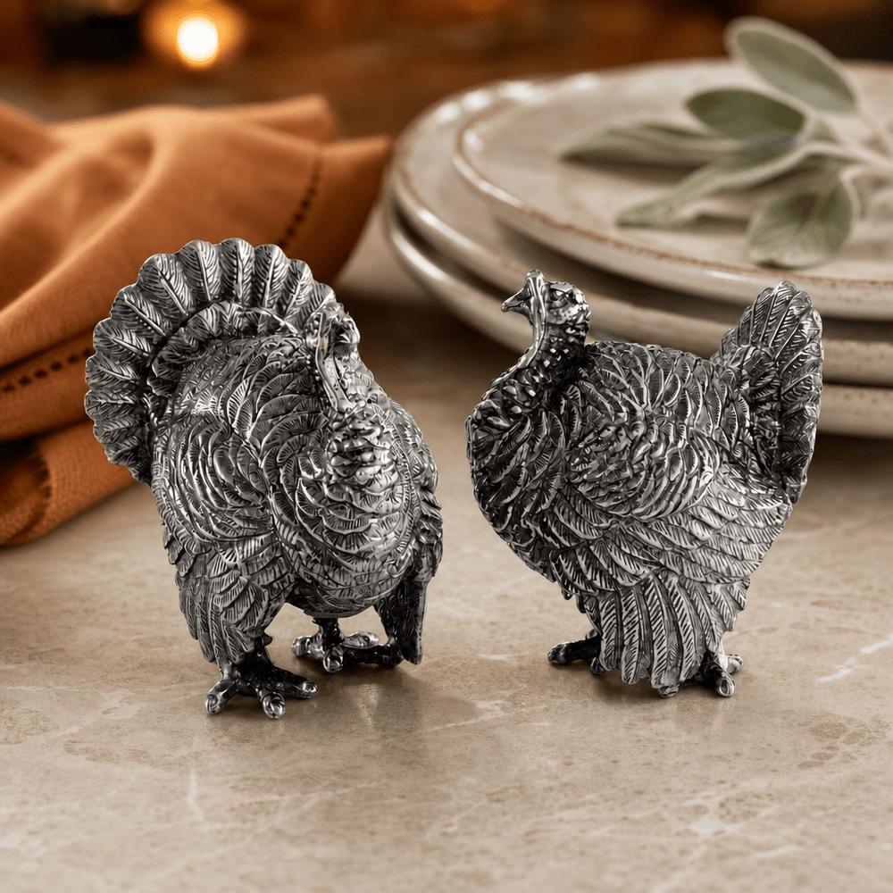 Pewter turkey salt and pepper shakers on a marble table beside stacked plates and a rust napkin