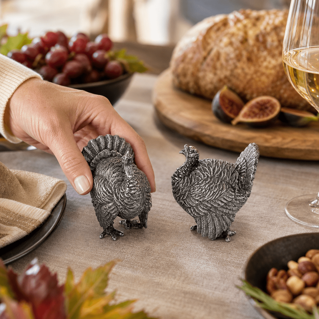 Hand lifting a pewter turkey shaker from a Thanksgiving table with bread, figs, and grapes