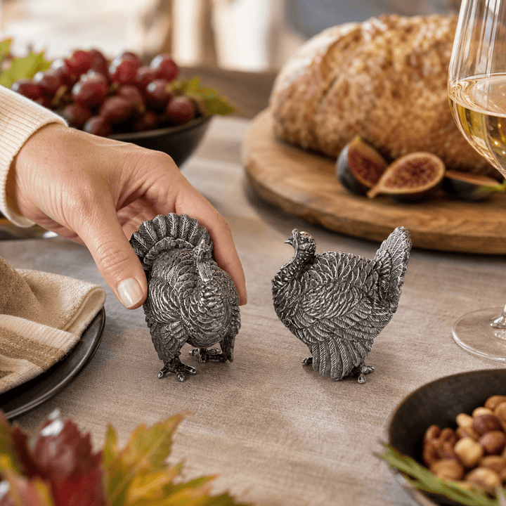 Hand lifting a pewter turkey shaker from a Thanksgiving table with bread, figs, and grapes