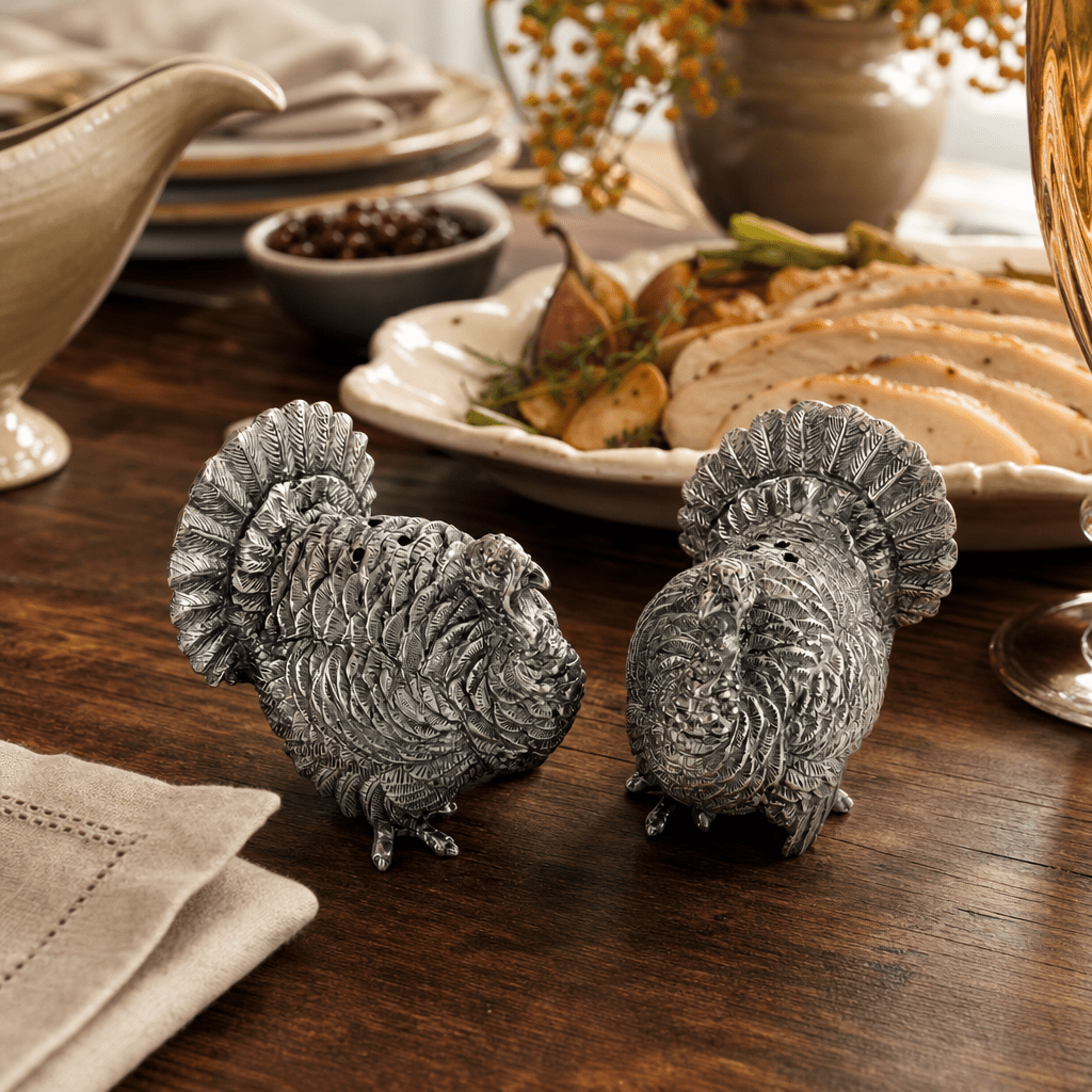 Pewter turkey salt and pepper shakers on a wood table beside sliced roast turkey and a gravy boat