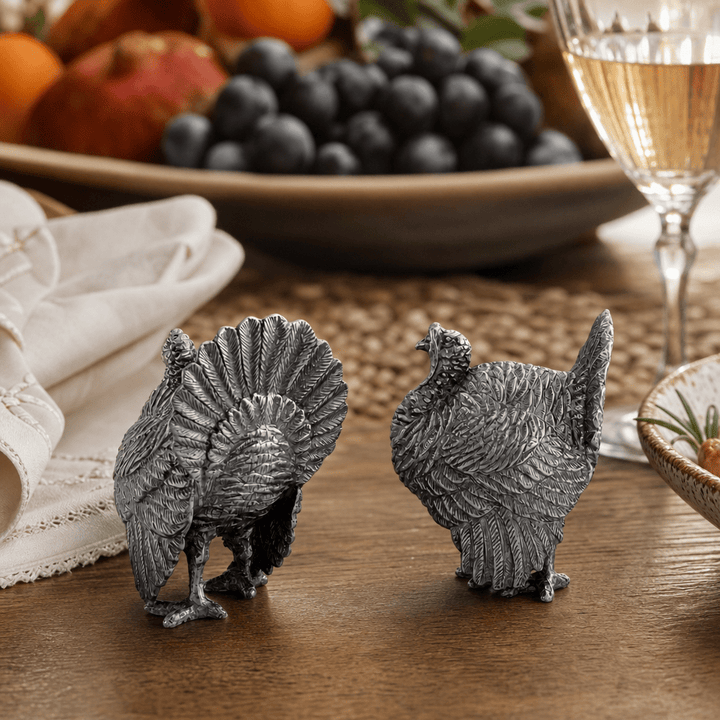 Pewter turkey salt and pepper shakers on a wood table with grapes, oranges, and a wine glass