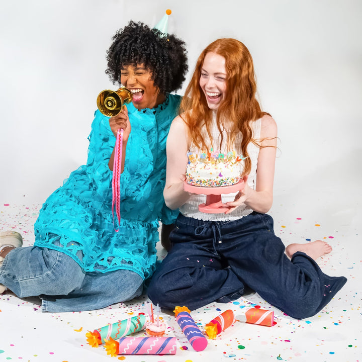 Two friends laughing on a confetti-covered floor, one shouting into the gold megaphone