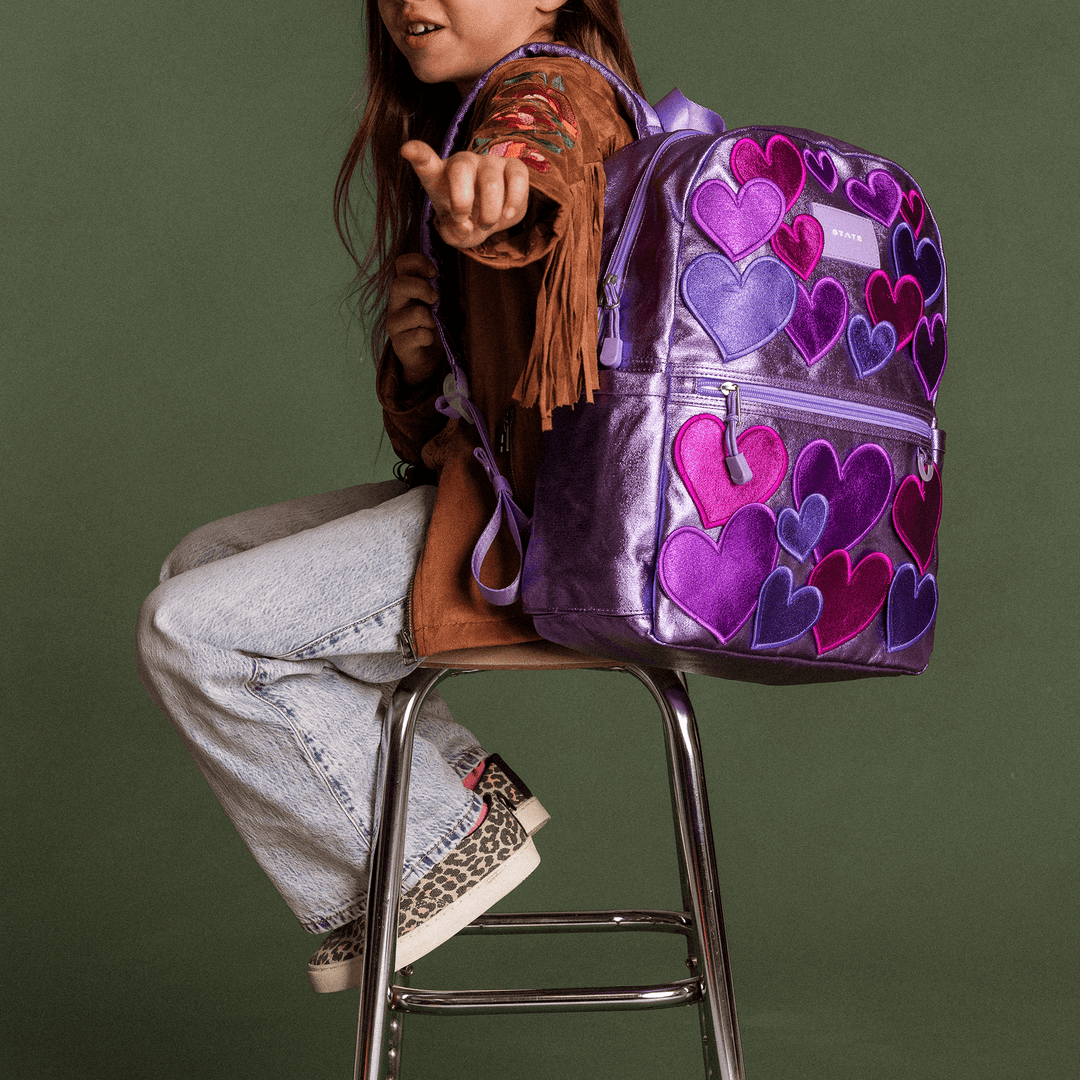 Girl sitting on a stool holding the lilac 3D Hearts STATE Kane backpack on her lap