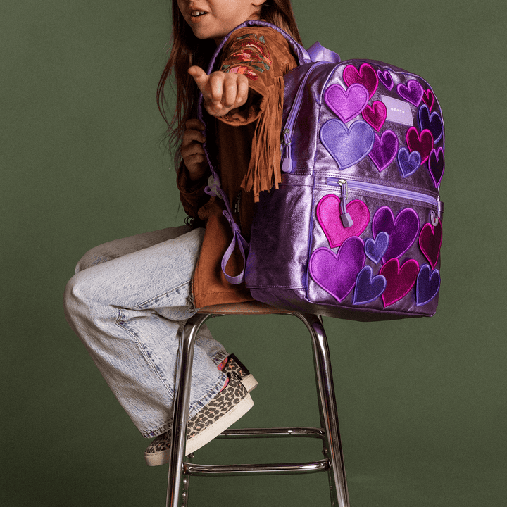 Girl sitting on a stool holding the lilac 3D Hearts STATE Kane backpack on her lap