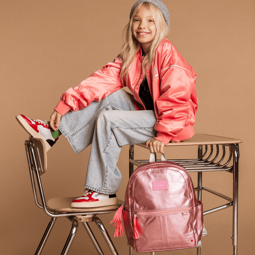 Girl in a pink bomber jacket sitting on a desk with the metallic pink STATE Kane backpack