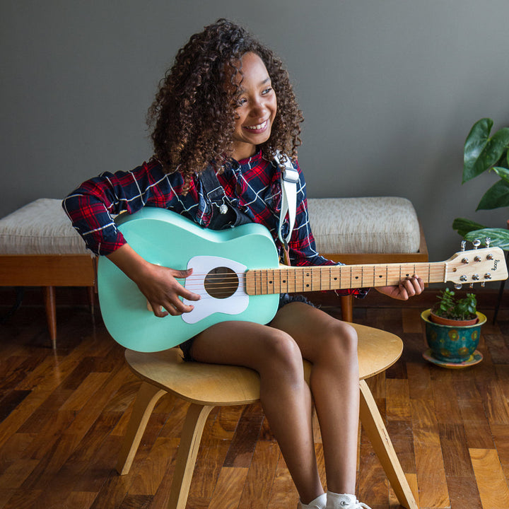 Woman holding a turquoise guitar in a room with wooden floor and gray walls