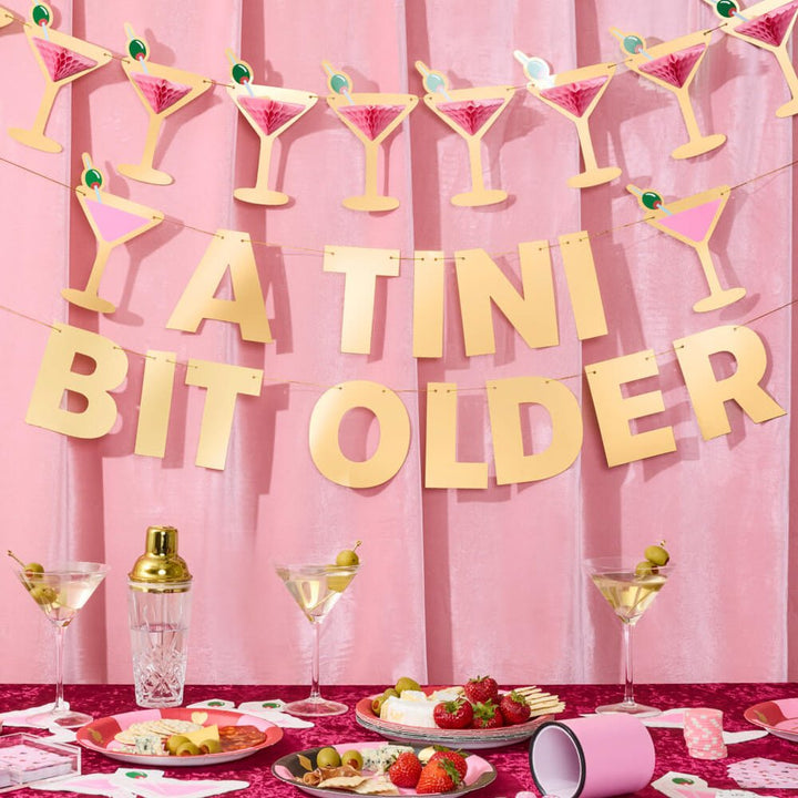 A Tini Bit Older birthday banner hung under a pink martini glass honeycomb banner above a cocktail party table