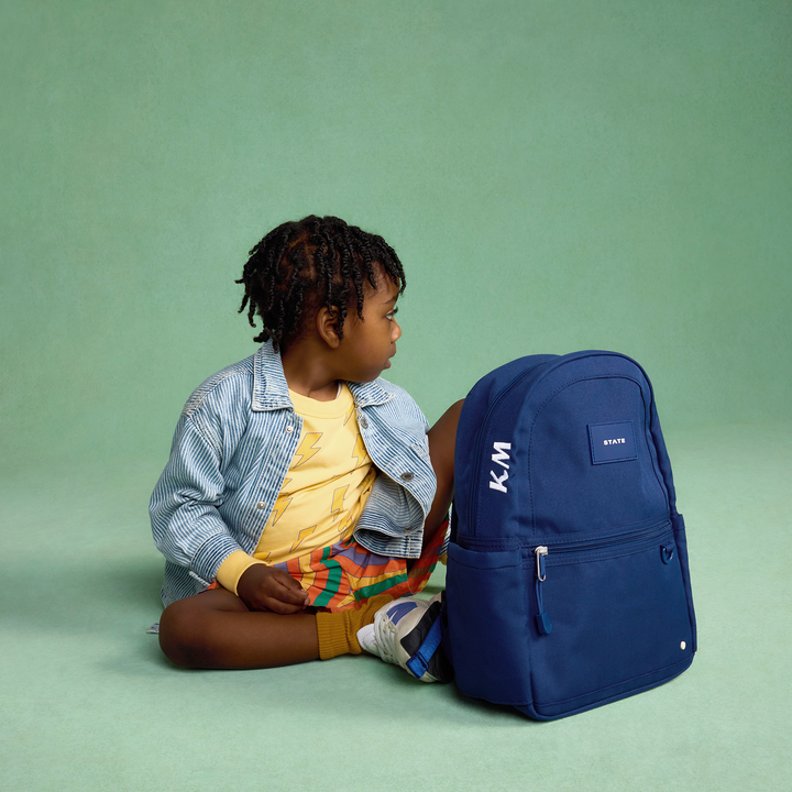 Toddler seated next to a navy Kane Mini backpack on a green backdrop