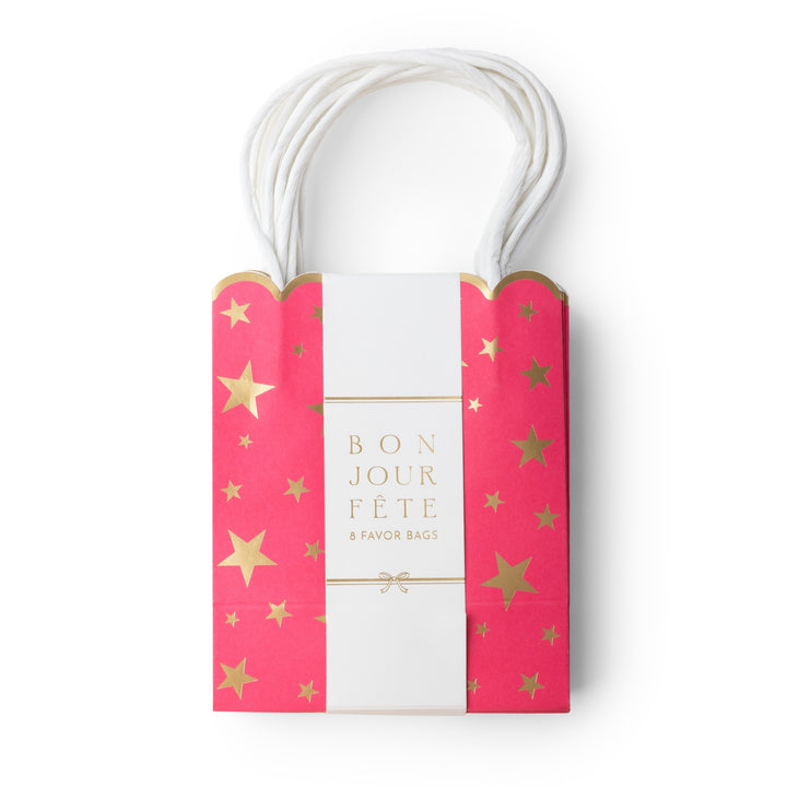 A pink paper gift bag with white handles, decorated with gold foil stars and a central white band reading 'BON JOUR FÈTE & FAVOR BAGS'.