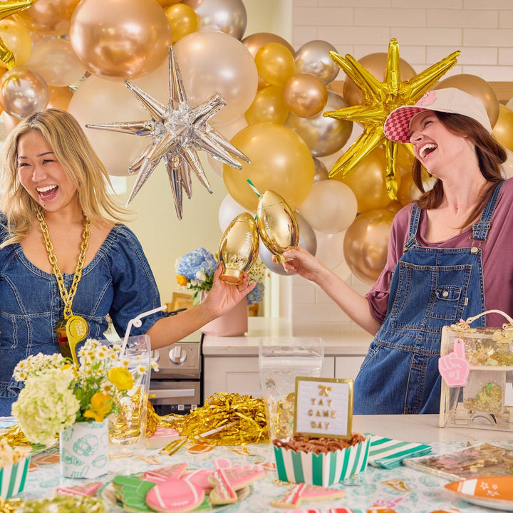 Two friends laughing at a gold-balloon game day party, one holding a gold football cup