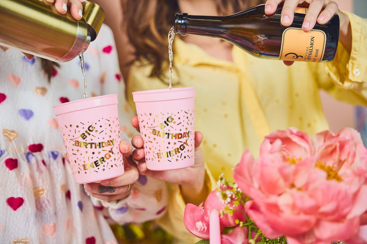 Two friends pouring bubbly into pink Big Birthday Energy cups beside pink peonies