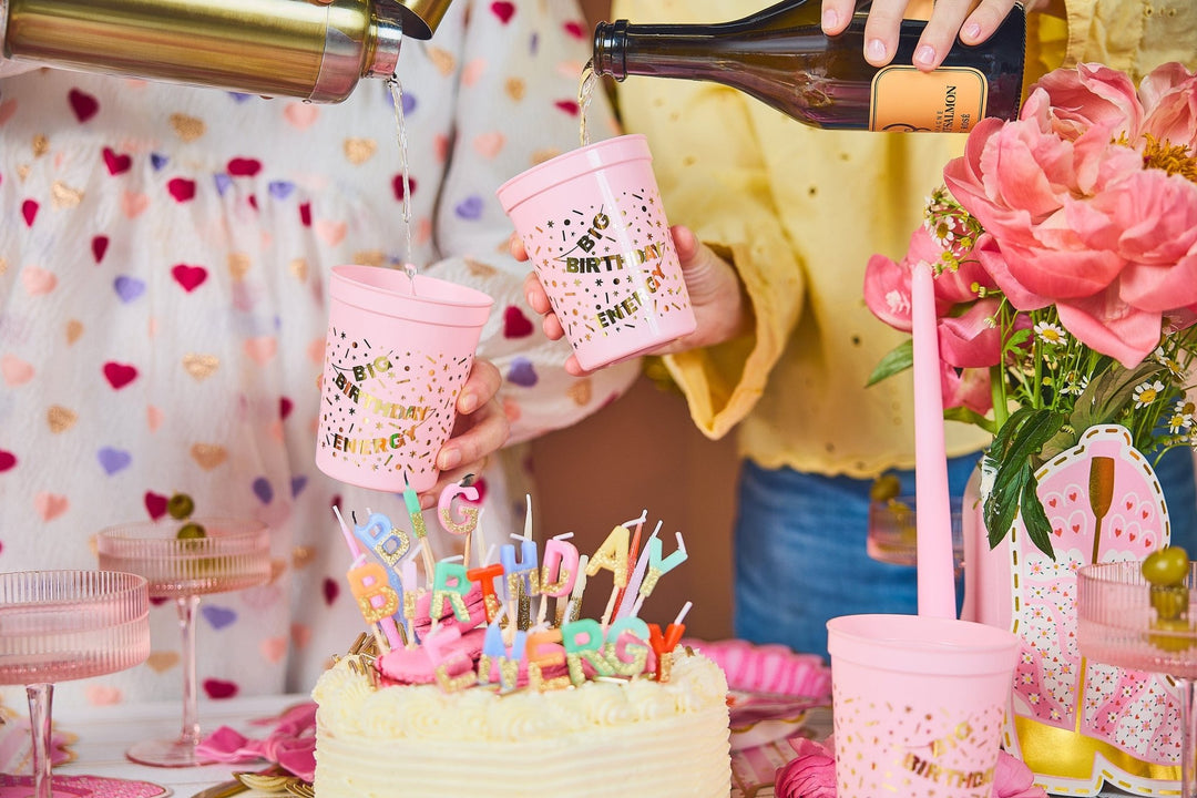 Pink Big Birthday Energy cups raised over a birthday cake with candle letters and peonies