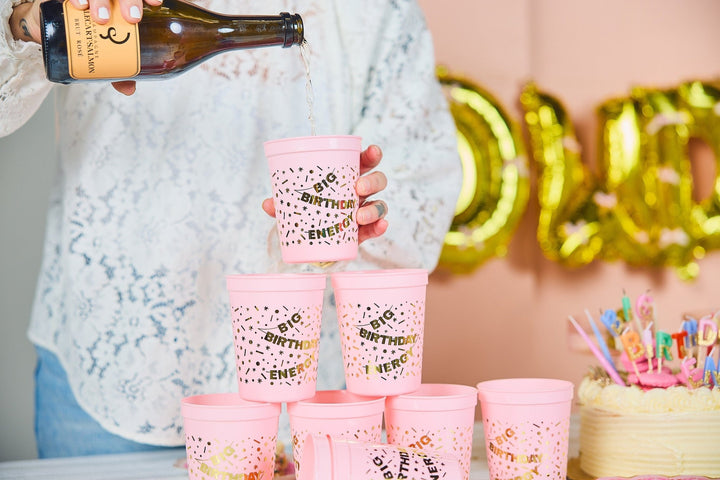 Pink birthday cups stacked into a pyramid in front of gold letter balloons and a cake
