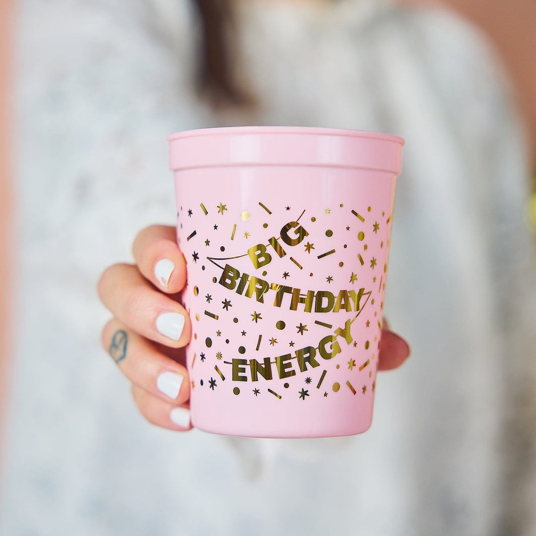 Hand holding a pink reusable cup printed Big Birthday Energy in gold foil lettering