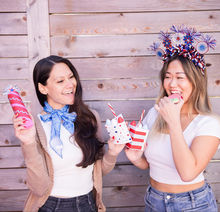 Two women at a 4th of July party holding cake slice cups and wearing firework headbands