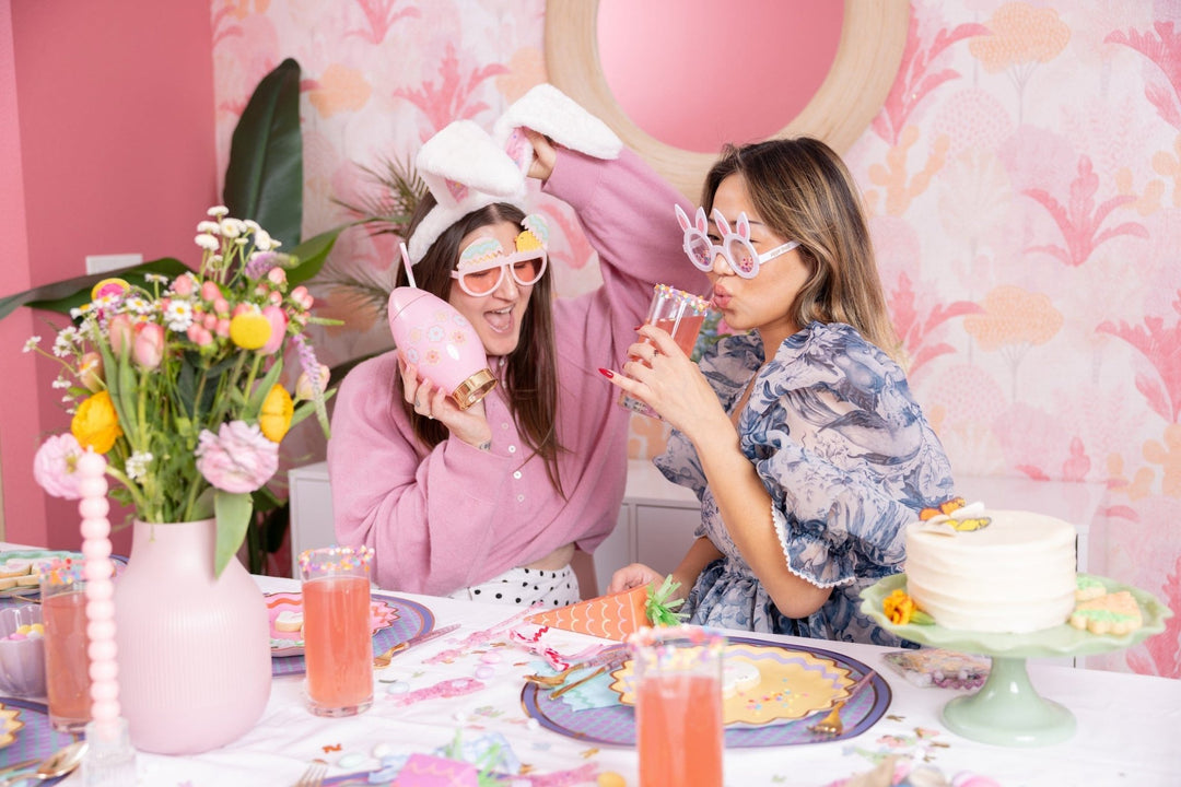 Two friends sipping pink drinks at an Easter table, one in the confetti bunny ears