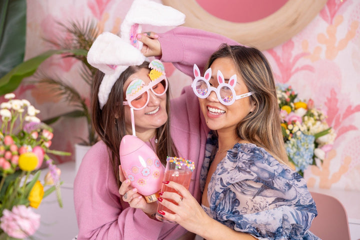 Two friends in bunny ears and bunny glasses posing with a pink Easter egg cup