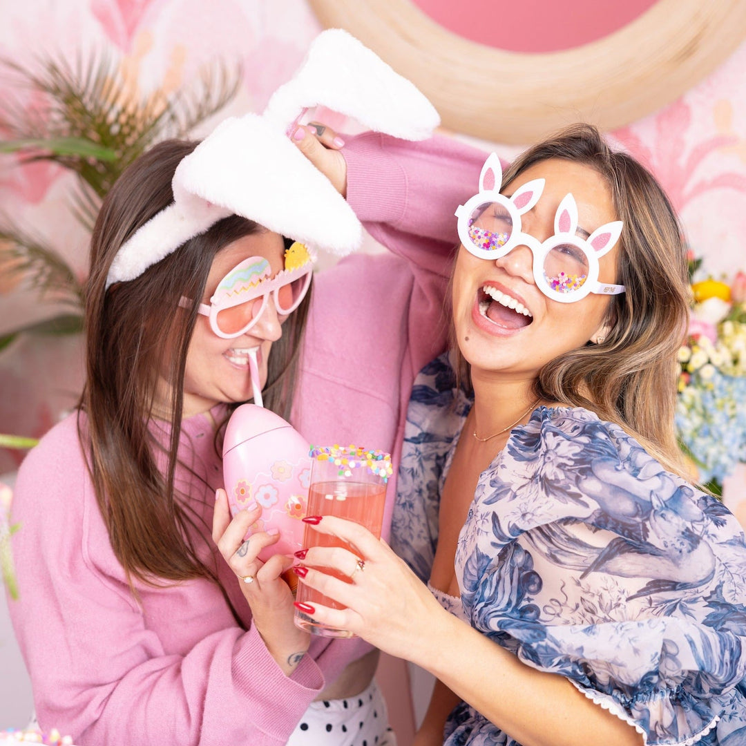 Two friends laughing in bunny ears and glasses with pink drinks at an Easter party