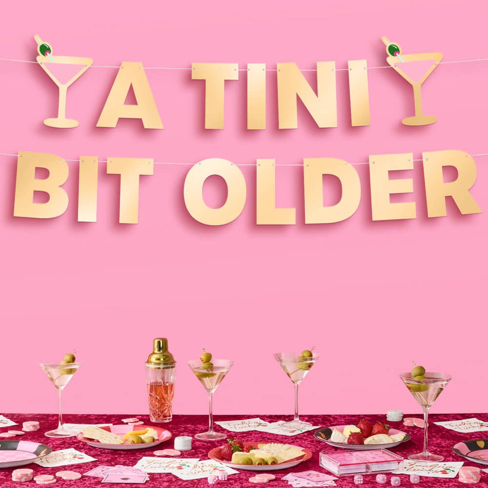 Gold foil A Tini Bit Older birthday banner on a pink wall over a cocktail party table set with martinis and snacks