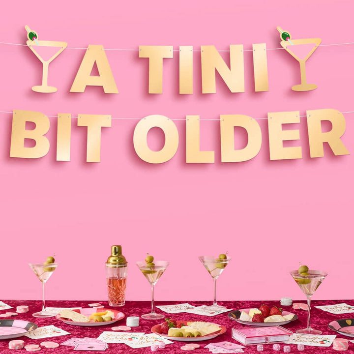 Gold foil A Tini Bit Older birthday banner on a pink wall over a cocktail party table set with martinis and snacks