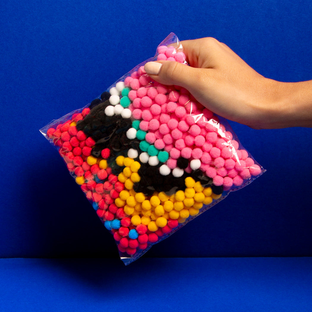 Hand holding a bag of 500-plus colorful pom poms on a blue background