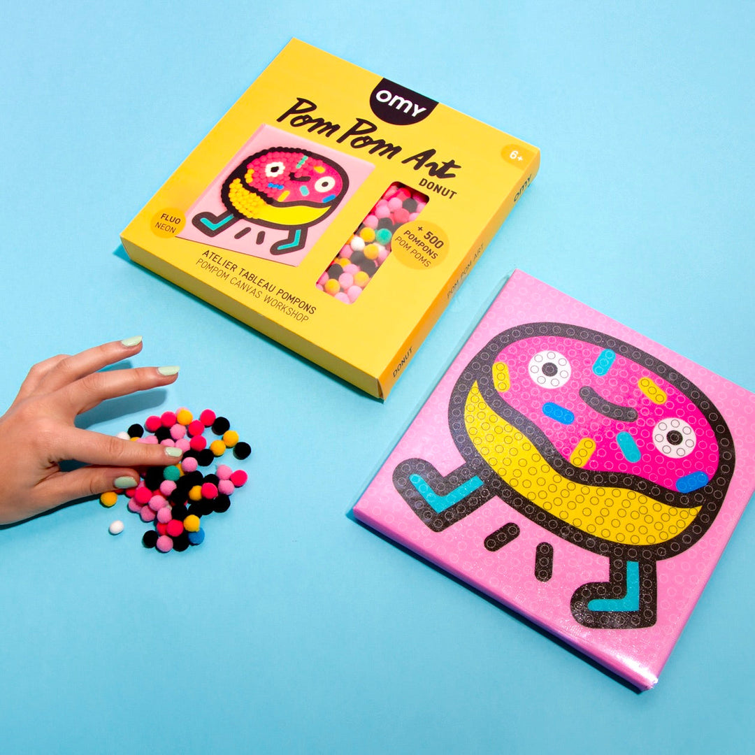 Donut pom pom art boards, one blank and one finished, with loose pompoms and hands, on blue