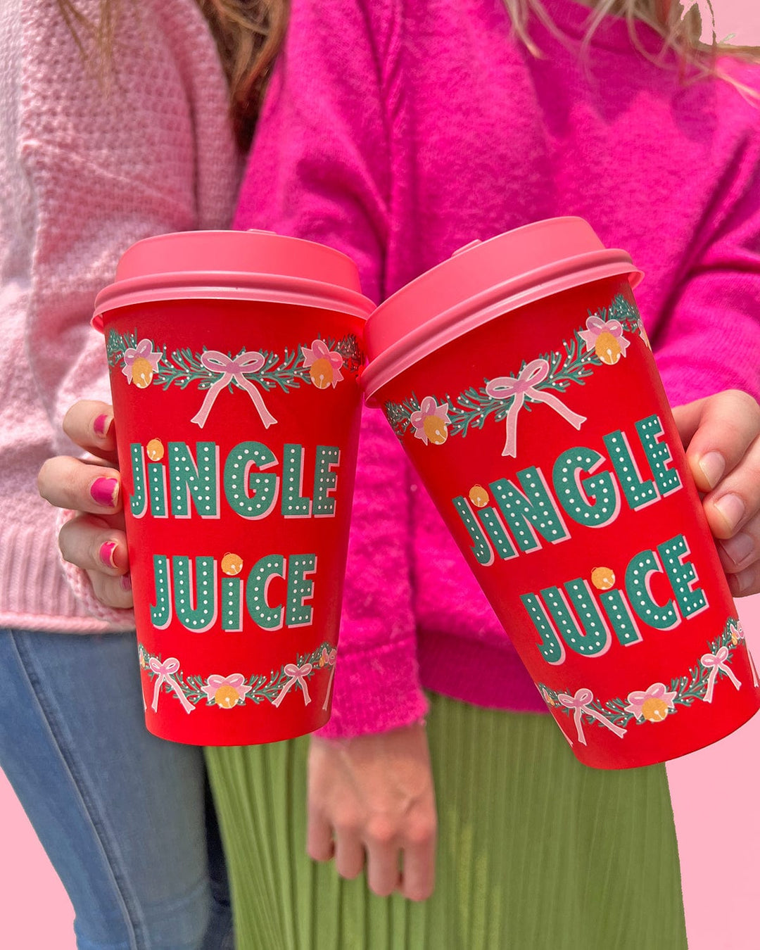 Hands holding red Jingle Juice Christmas to-go cups against a pink background