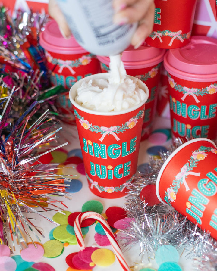 Hand adding whipped cream to a red Jingle Juice Christmas cup surrounded by confetti