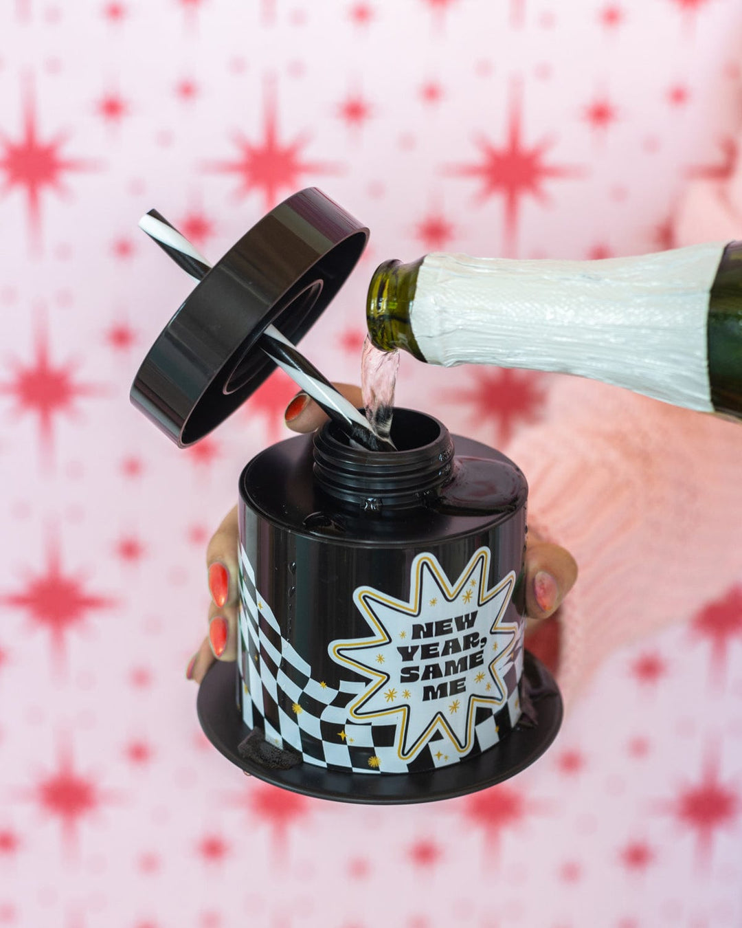 Champagne poured into the open New Year, Same Me top hat cup against a pink star backdrop