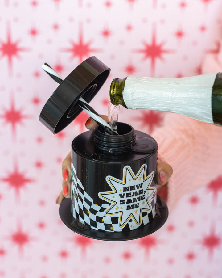 Champagne poured into the open New Year, Same Me top hat cup against a pink star backdrop