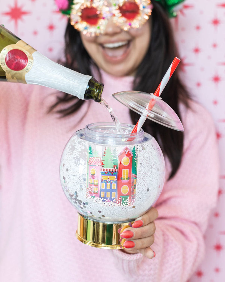 Woman in festive glasses pouring champagne into a snow globe sipper cup against a pink background