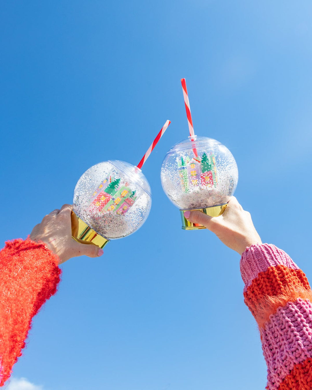 Two hands raising holiday village snow globe cups against a bright blue sky