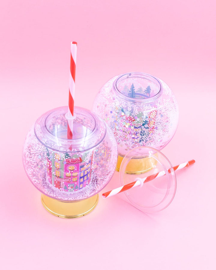 Two holiday village snow globe cups with gold bases and striped straws on a pink background