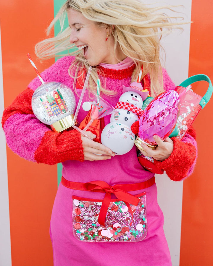 Woman in a pink sweater holding the snow globe cup and a snowman sipper in front of an orange wall