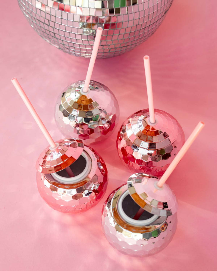 Silver and pink disco ball cups with straws under a large mirror ball on a pink table