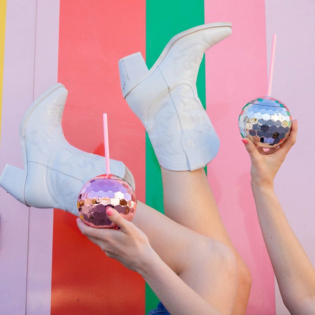 Two raised legs in white cowboy boots and hands holding pink and silver disco ball cups against stripes