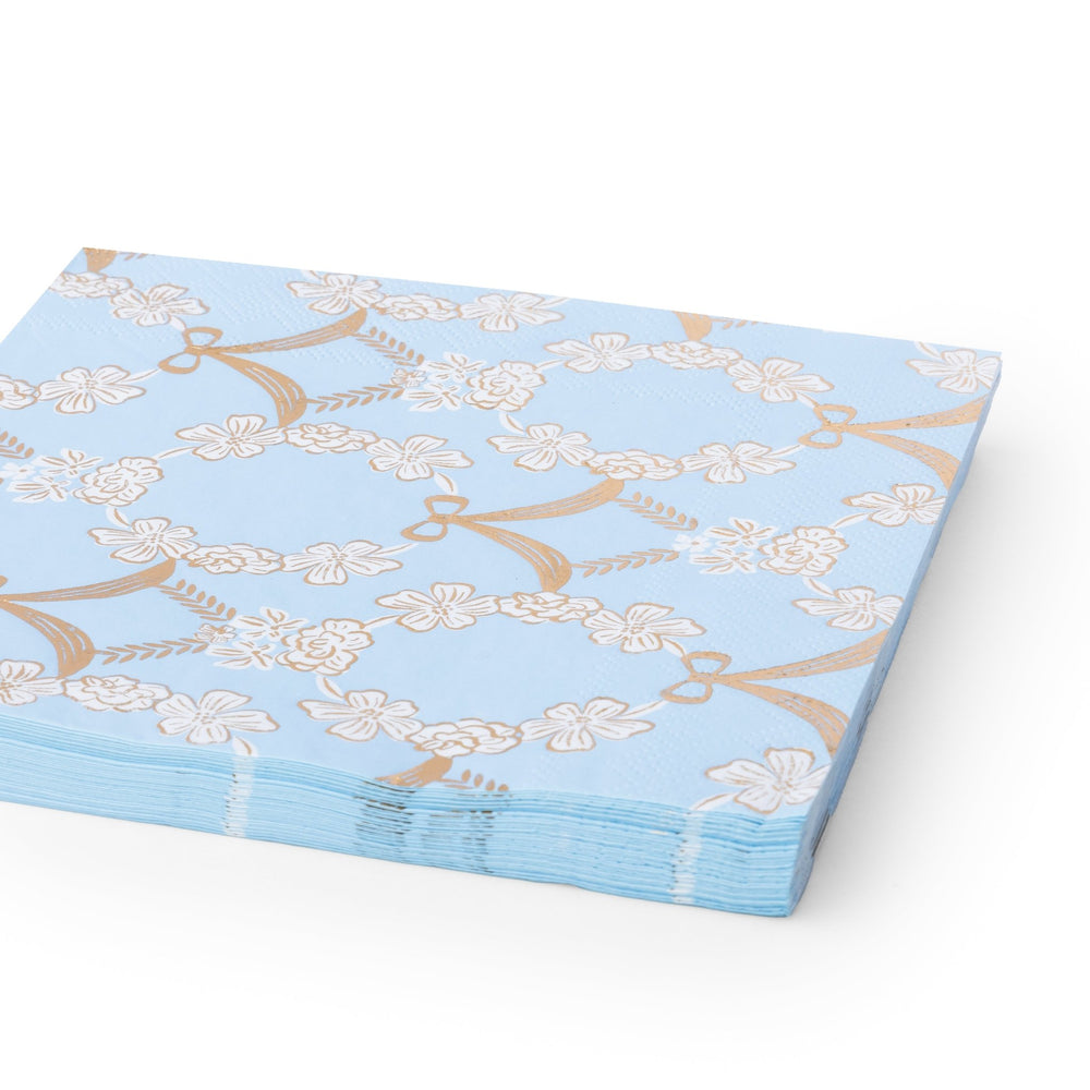 A stack of light blue paper napkins featuring a repeating pattern of white flowers and gold ribbons, shown on a clean white background.