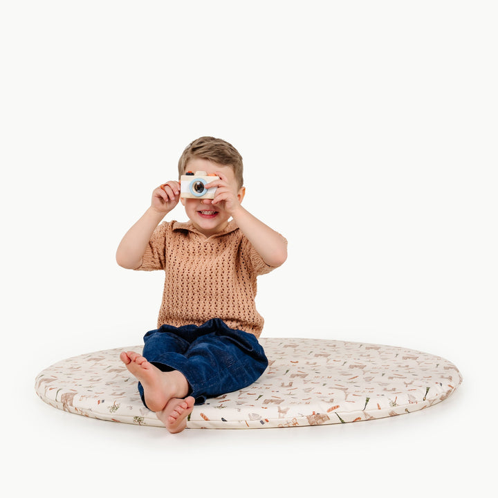 Boy taking a photo with a wooden toy camera while sitting on a round cream padded play mat