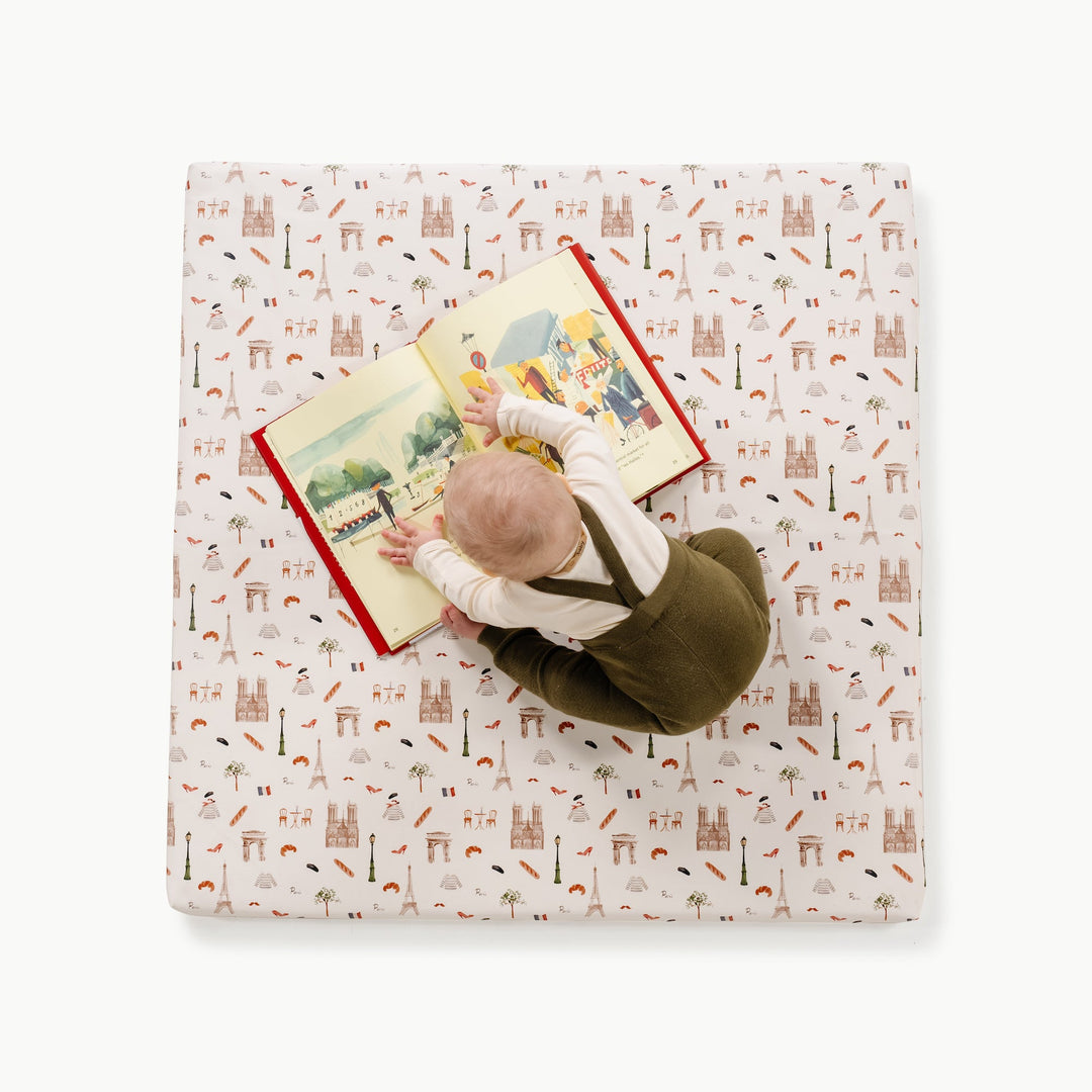 Overhead view of a baby reading a book on the square Paris print padded play mat with Paris landmark motifs