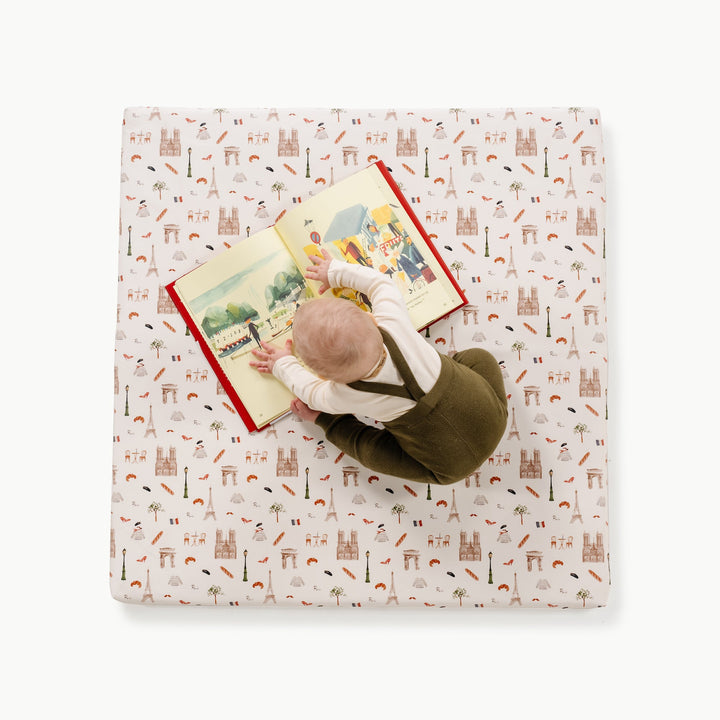 Overhead view of a baby reading a book on the square Paris print padded play mat with Paris landmark motifs