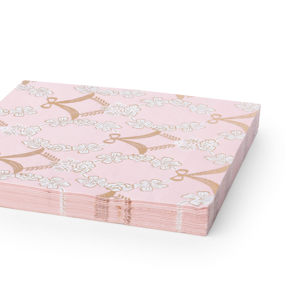 A stack of pink paper napkins featuring a repeating pattern of white cherry blossoms and gold bow motifs, shown on a clean white background.