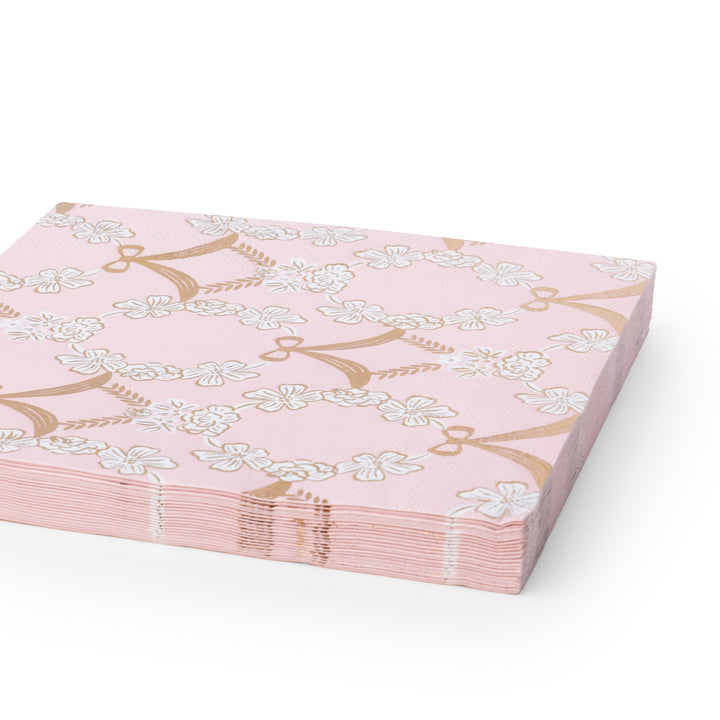 A stack of pink paper napkins featuring a repeating pattern of white cherry blossoms and gold bow motifs, shown on a clean white background.