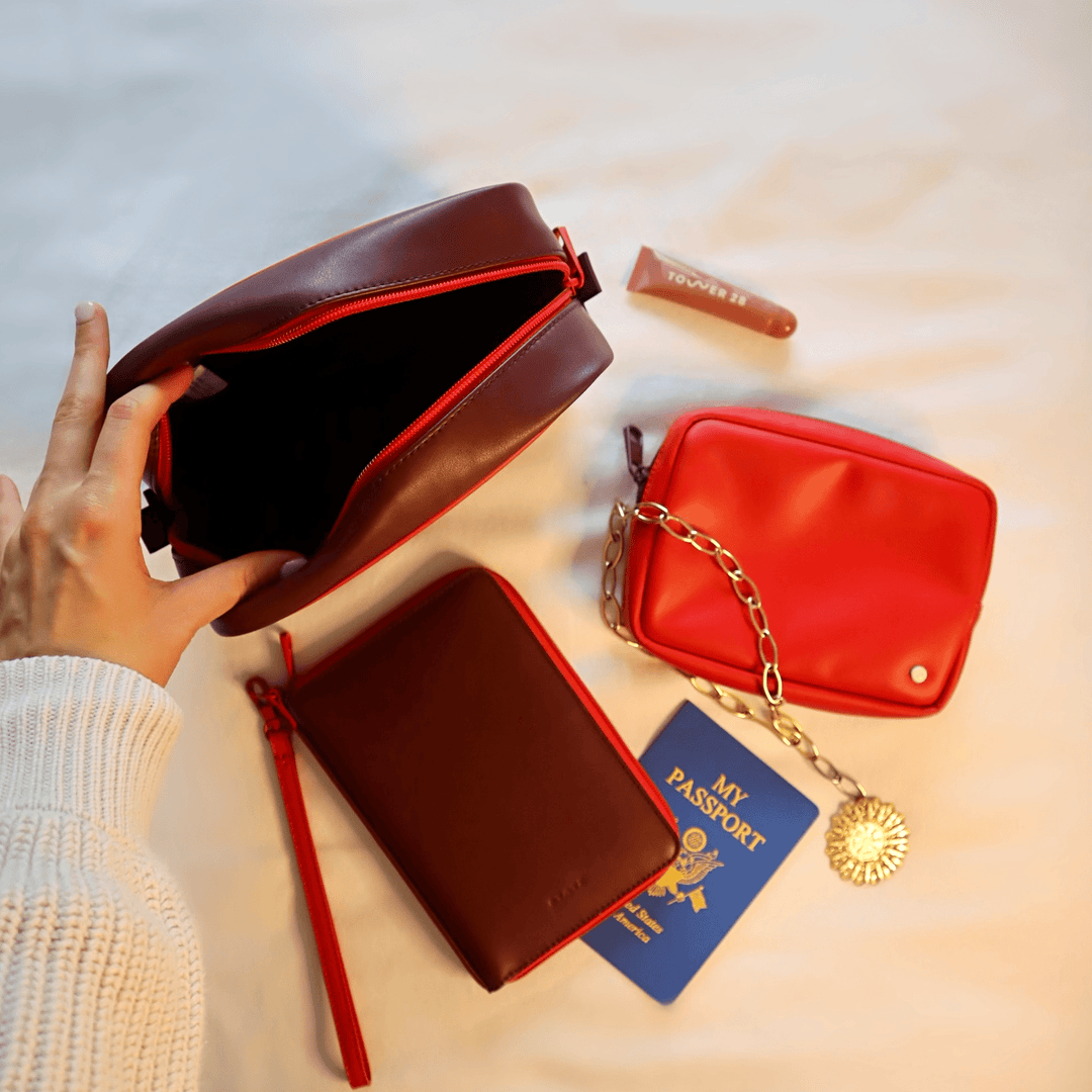 Burgundy and red pouches styled on a bed with a passport, lip gloss, and a gold chain
